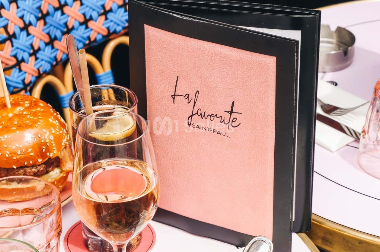 Menu rose posé sur une table avec un verre de vin rosé, un burger et des couverts en terrasse.