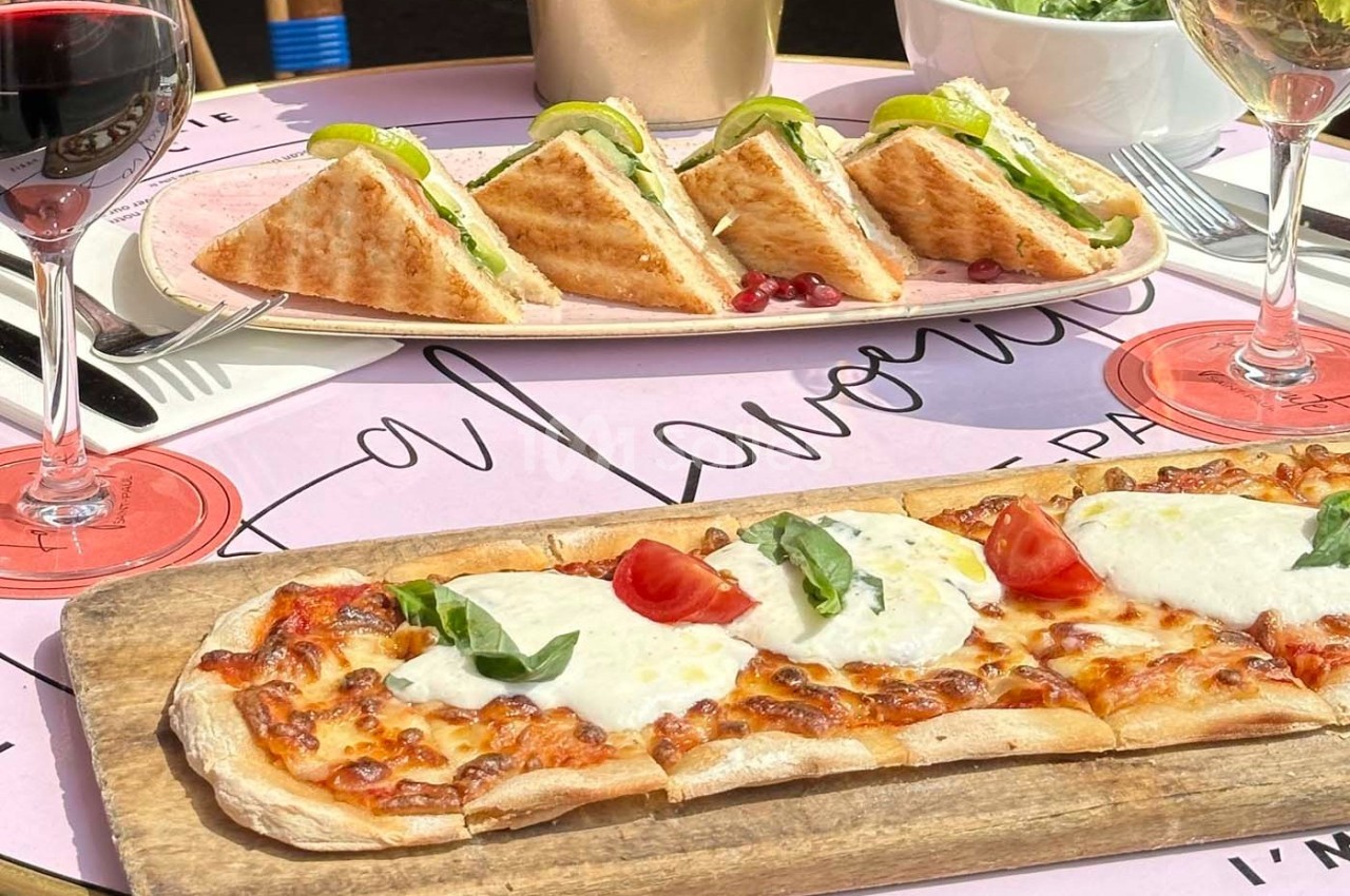 Assiette de sandwichs triangulaires et planche de pizza garnie de mozzarella et tomates, avec verres de vin rouge.