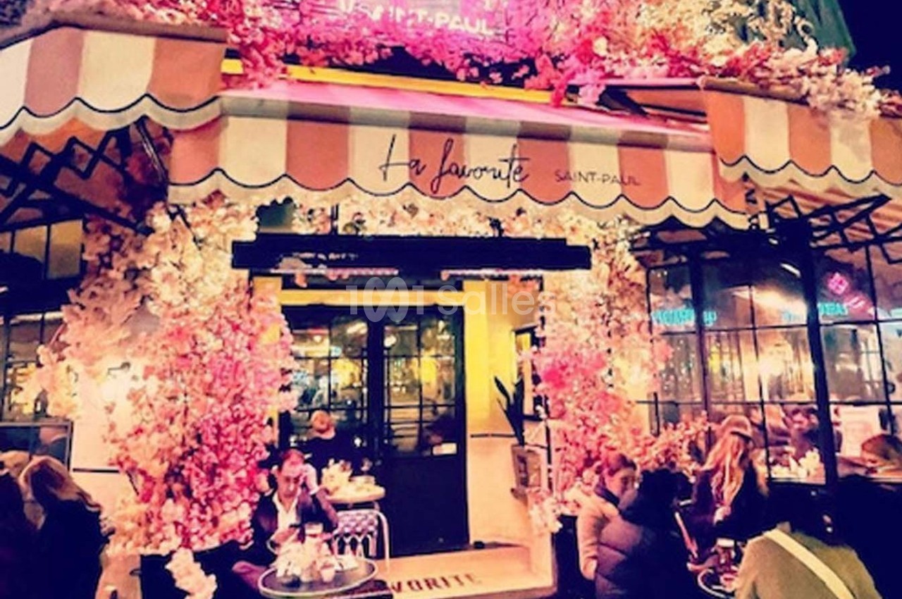 Façade d'un café décoré de fleurs roses avec des clients attablés en terrasse le soir.