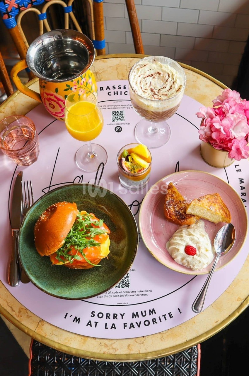 Table ronde avec un repas composé d'un burger, desserts, boissons et fleurs roses en décoration.
