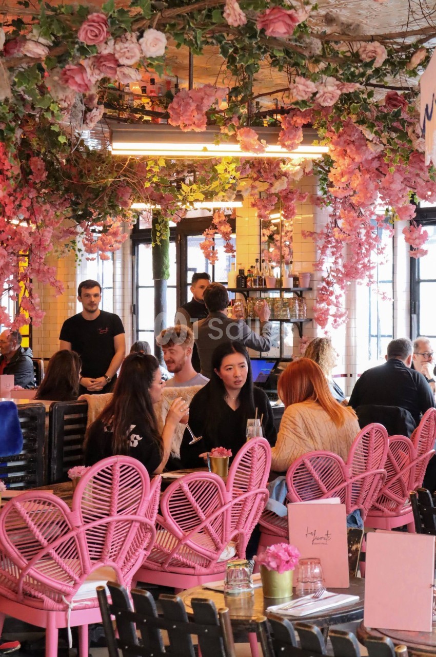 Salle de restaurant décorée de fleurs roses suspendues, avec des clients assis sur des chaises roses et noires.