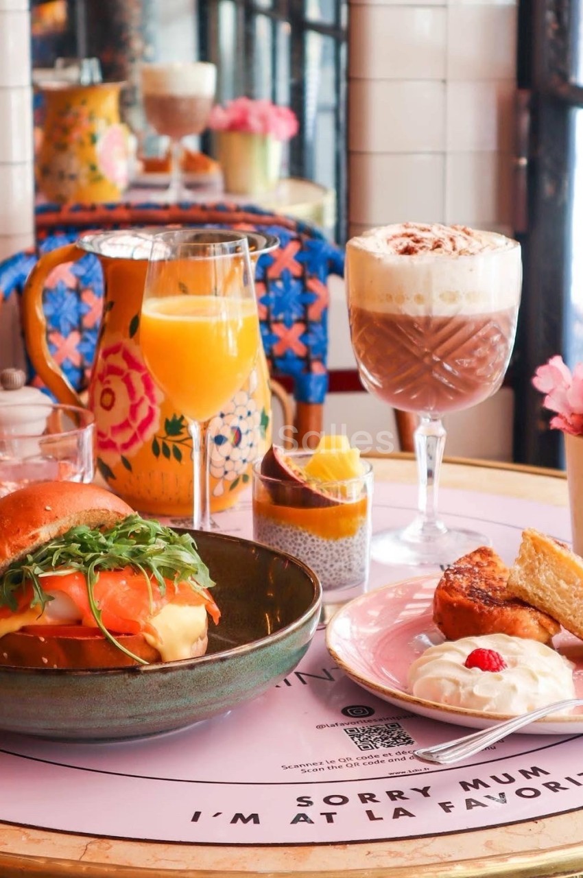 Petit-déjeuner composé d'un burger, d'un verre de jus, d'un chocolat chaud, de viennoiseries et de fruits sur une table…