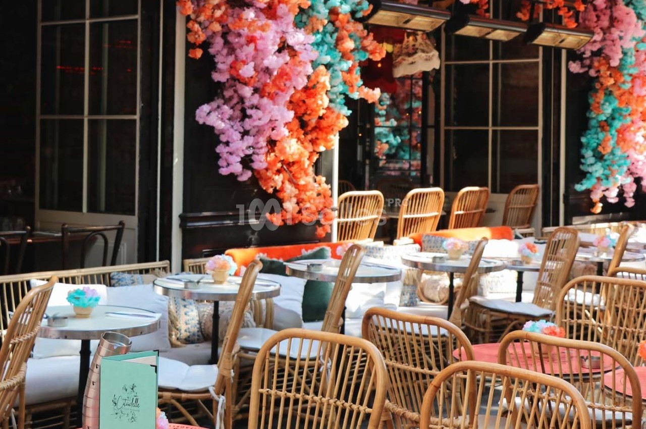 Terrasse d'un café avec des chaises en rotin, des tables rondes et des décorations florales colorées.