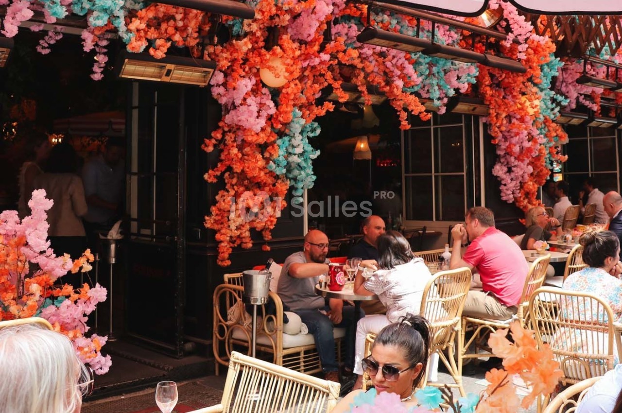 Terrasse de café animée avec des clients assis et une décoration florale colorée en orange et rose.