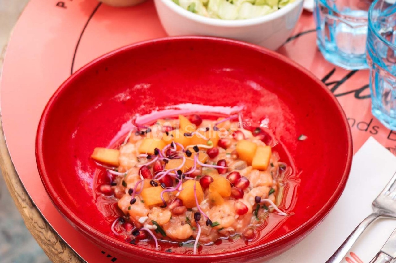 Bol rouge contenant une salade colorée avec morceaux de melon, graines de grenade et pousses, sur une table avec des verres.