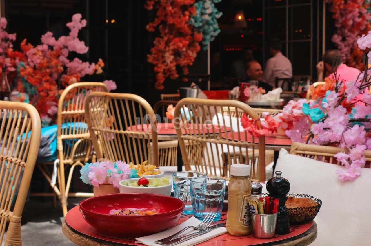 Table de restaurant en terrasse, décorée de fleurs colorées, avec des plats, condiments et verres disposés.