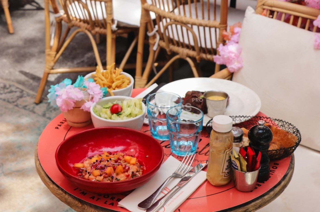Table ronde avec des plats variés, dont une salade colorée, des frites, et un steak, sur une terrasse ensoleillée.