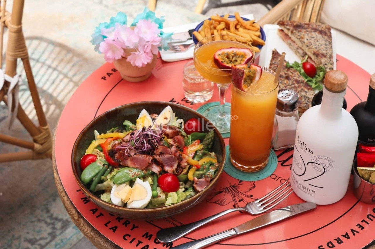 Assiette de salade garnie de légumes, œufs et viande, accompagnée de boissons, frites et pain sur une table colorée.