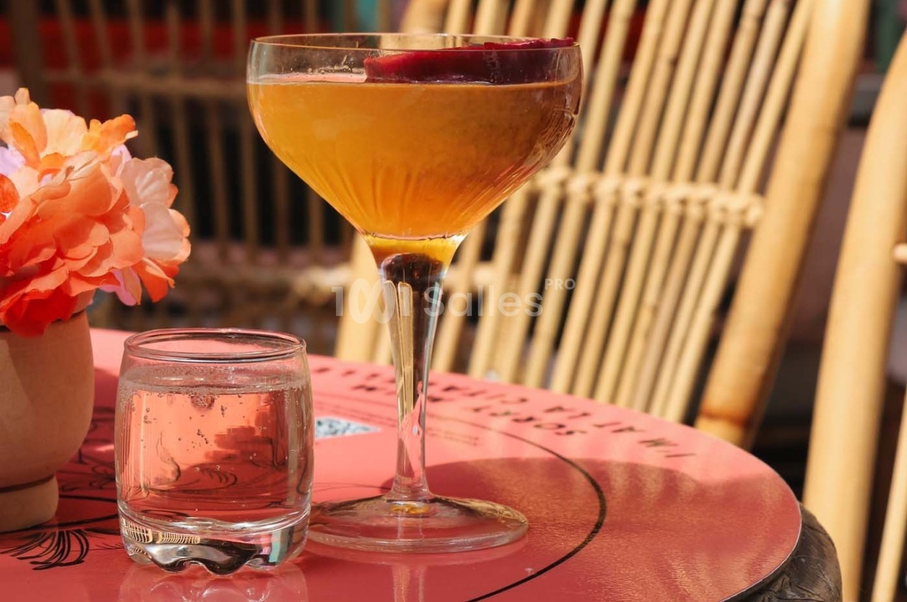 Verre à cocktail orange garni d'une tranche de fruit rouge, posé sur une table rose avec un verre d'eau et des fleurs.