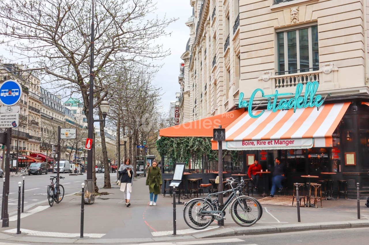Terrasse d’un café parisien avec auvent orange et blanc, située à l’angle d’une rue bordée d’arbres et de bâtiments.