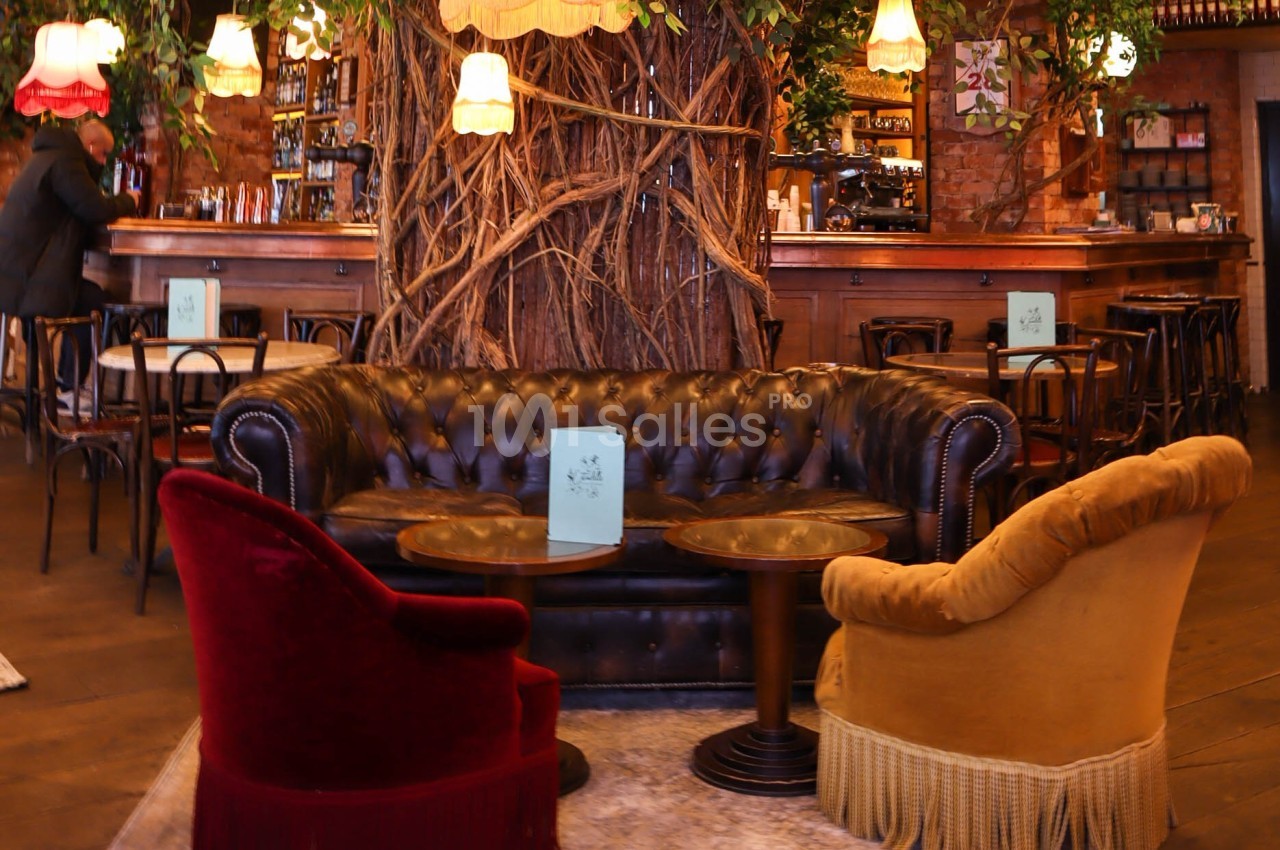 Salon cosy avec canapé en cuir, fauteuils colorés, tables basses et décor végétal dans un café-bar chaleureux.