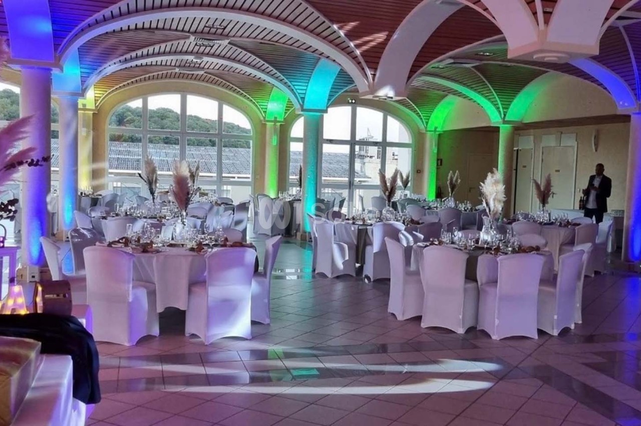 Salle de réception décorée avec des tables rondes, nappes blanches et éclairages colorés, vue sur l'extérieur.