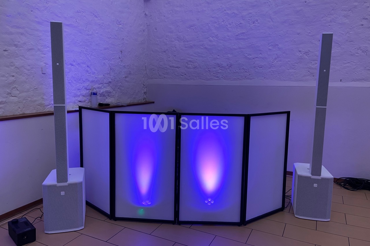 Deux enceintes blanches sur pied entourent une table de DJ éclairée par des lumières violettes dans une pièce aux murs…