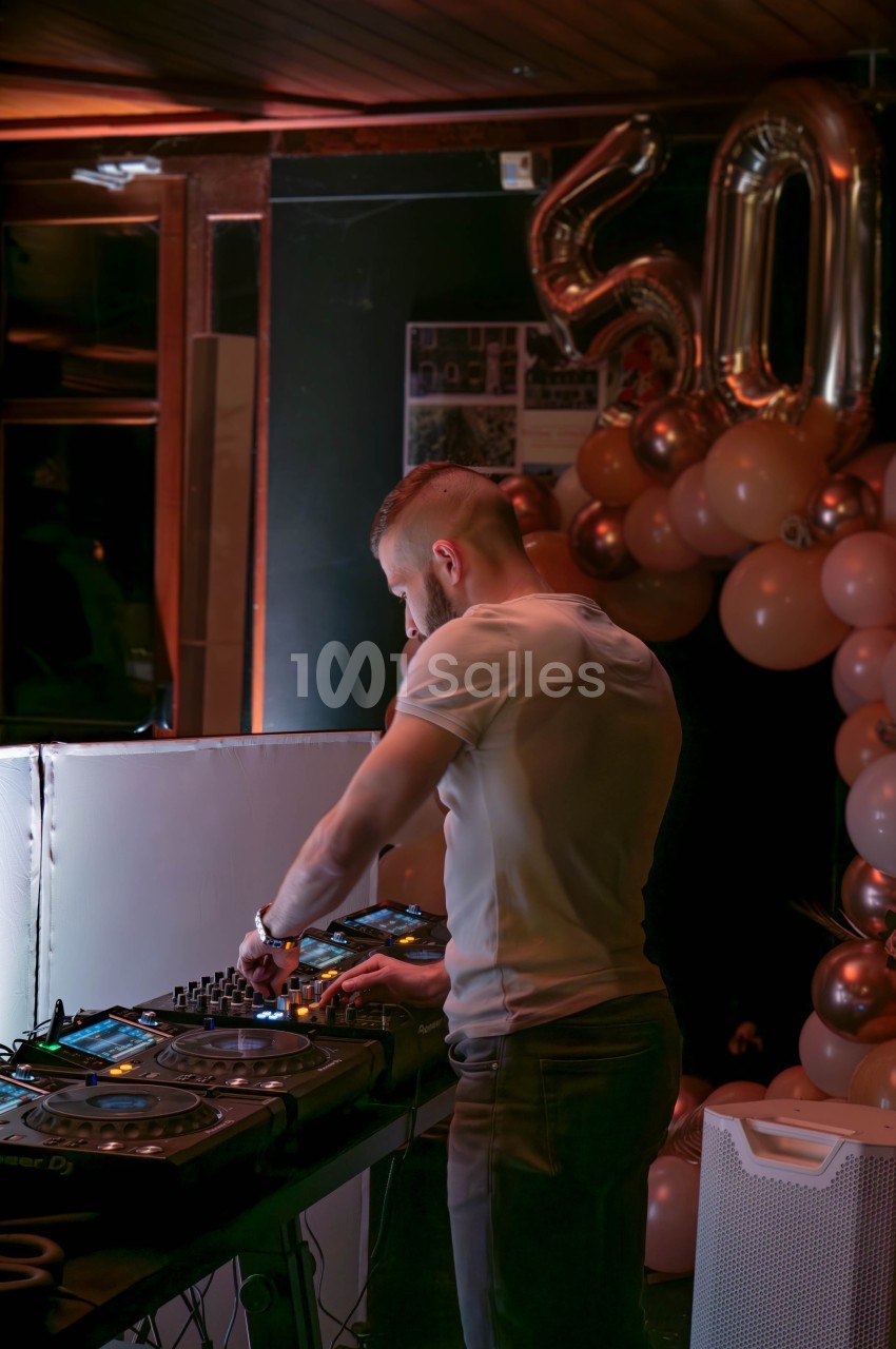 Un DJ mixe de la musique sur une table de mixage dans un décor festif avec des ballons et le chiffre 50 en arrière-plan.