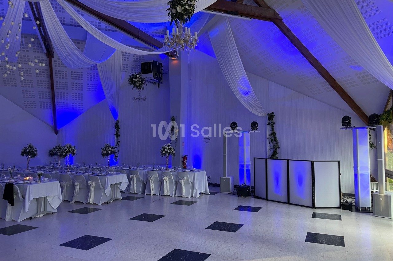 Salle de réception décorée avec des tables dressées, des drapés blancs suspendus et un éclairage bleu.