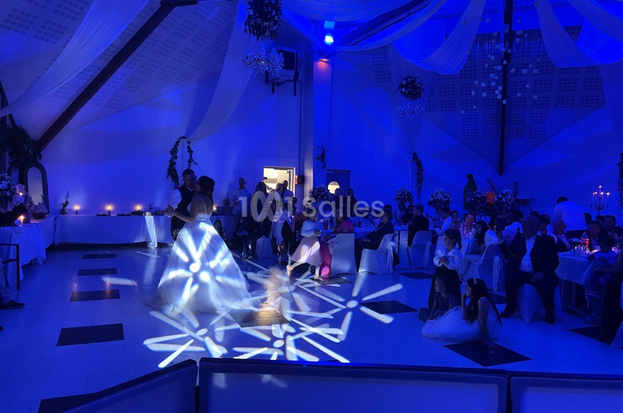 Un couple danse au centre d'une salle de réception éclairée en bleu, entouré d'invités assis.