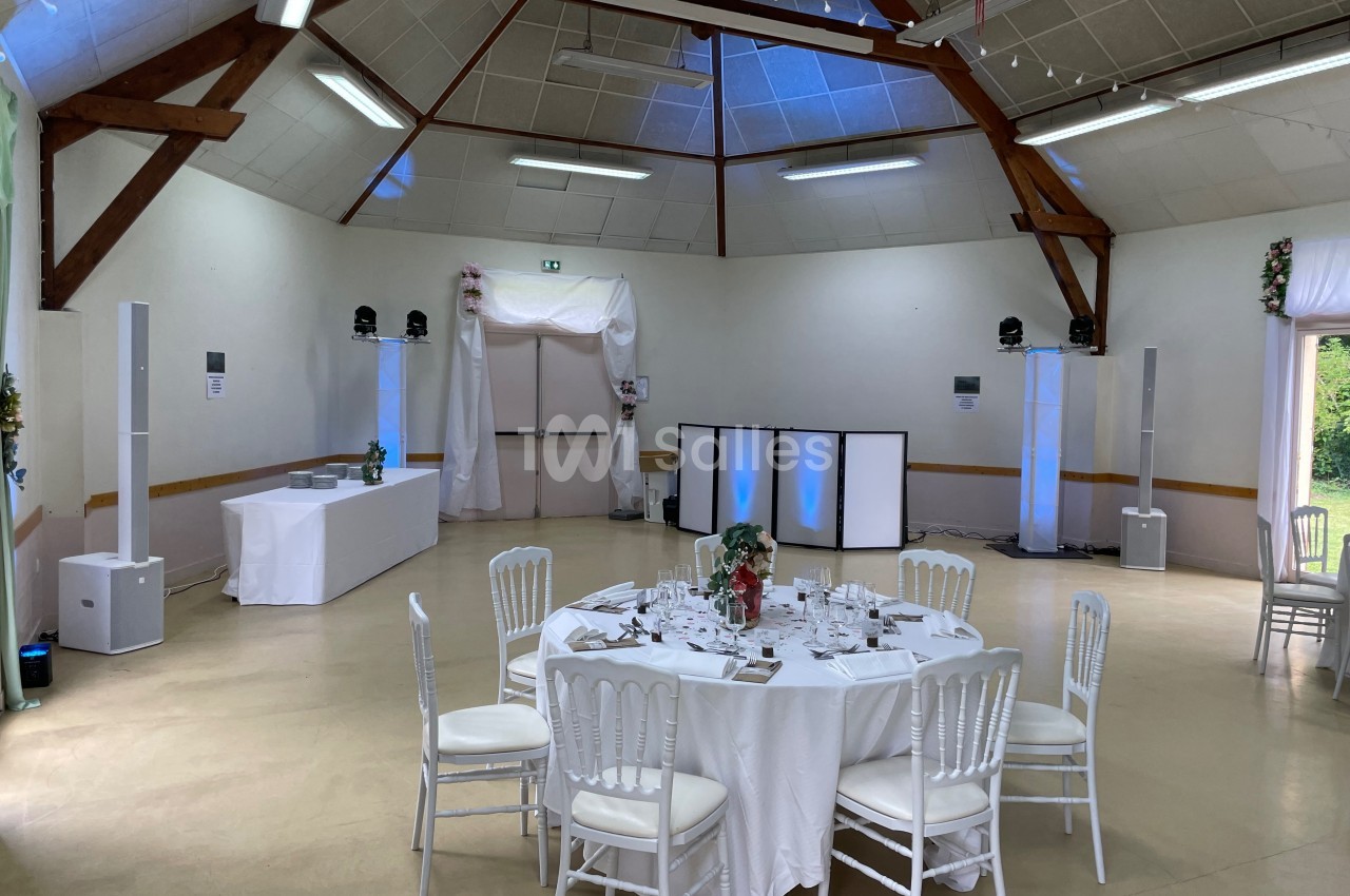 Salle de réception décorée avec des tables dressées, des chaises blanches et un éclairage discret.