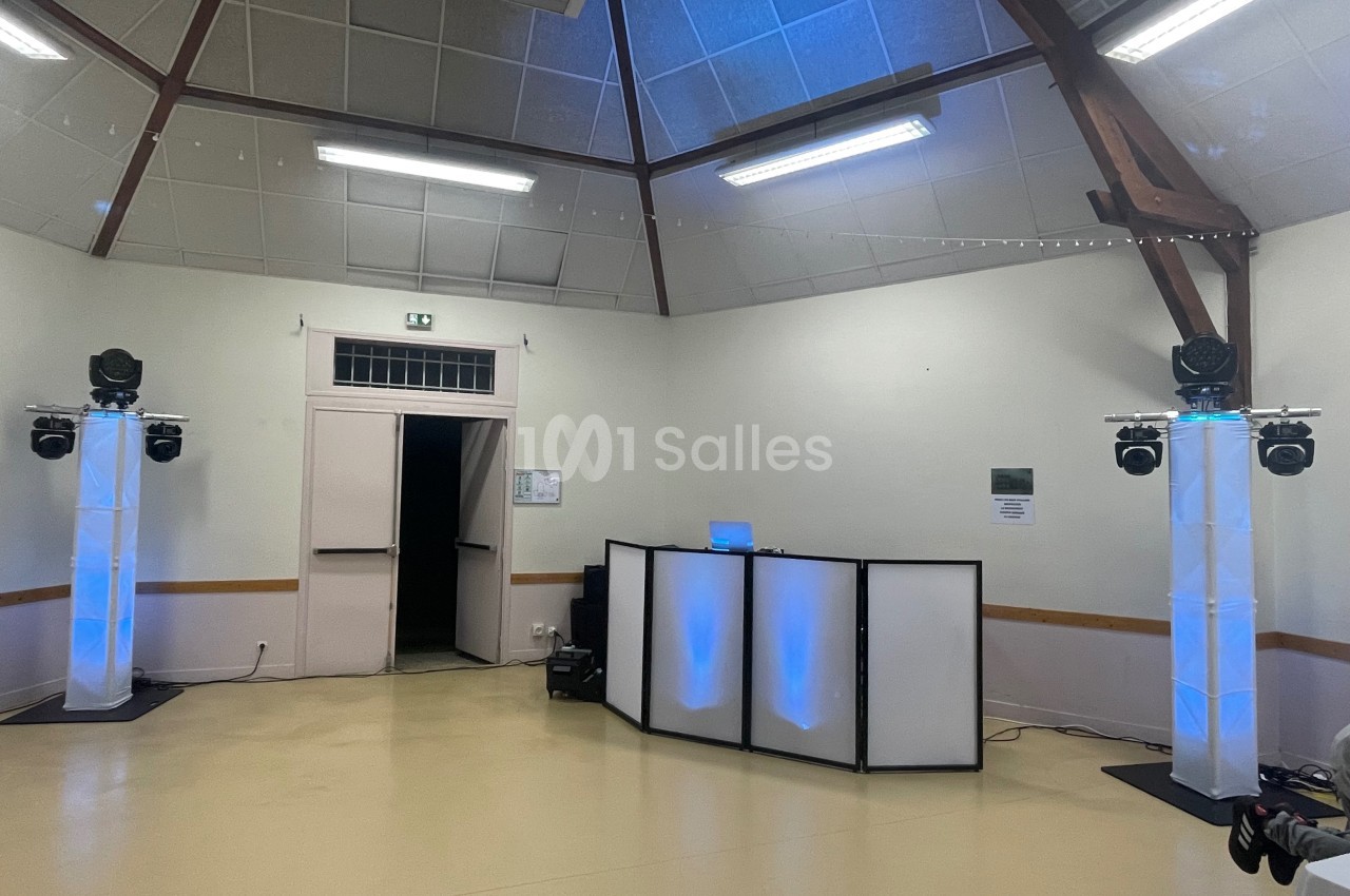Salle avec un espace DJ équipé de lumières colorées et d'enceintes, dans un décor sobre avec plafond en bois.