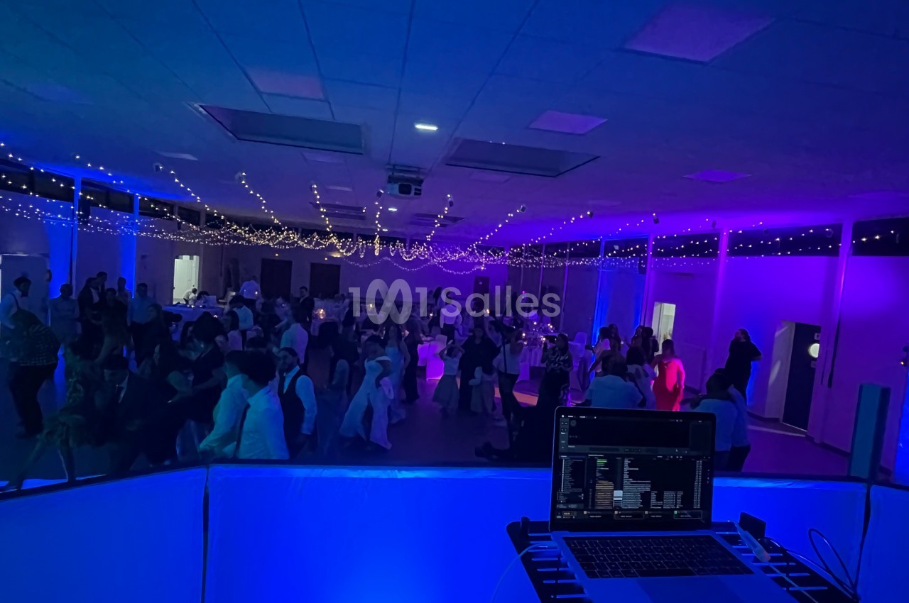 Salle de réception éclairée de lumières colorées avec des invités dansant, vue depuis une table de DJ équipée d'un…