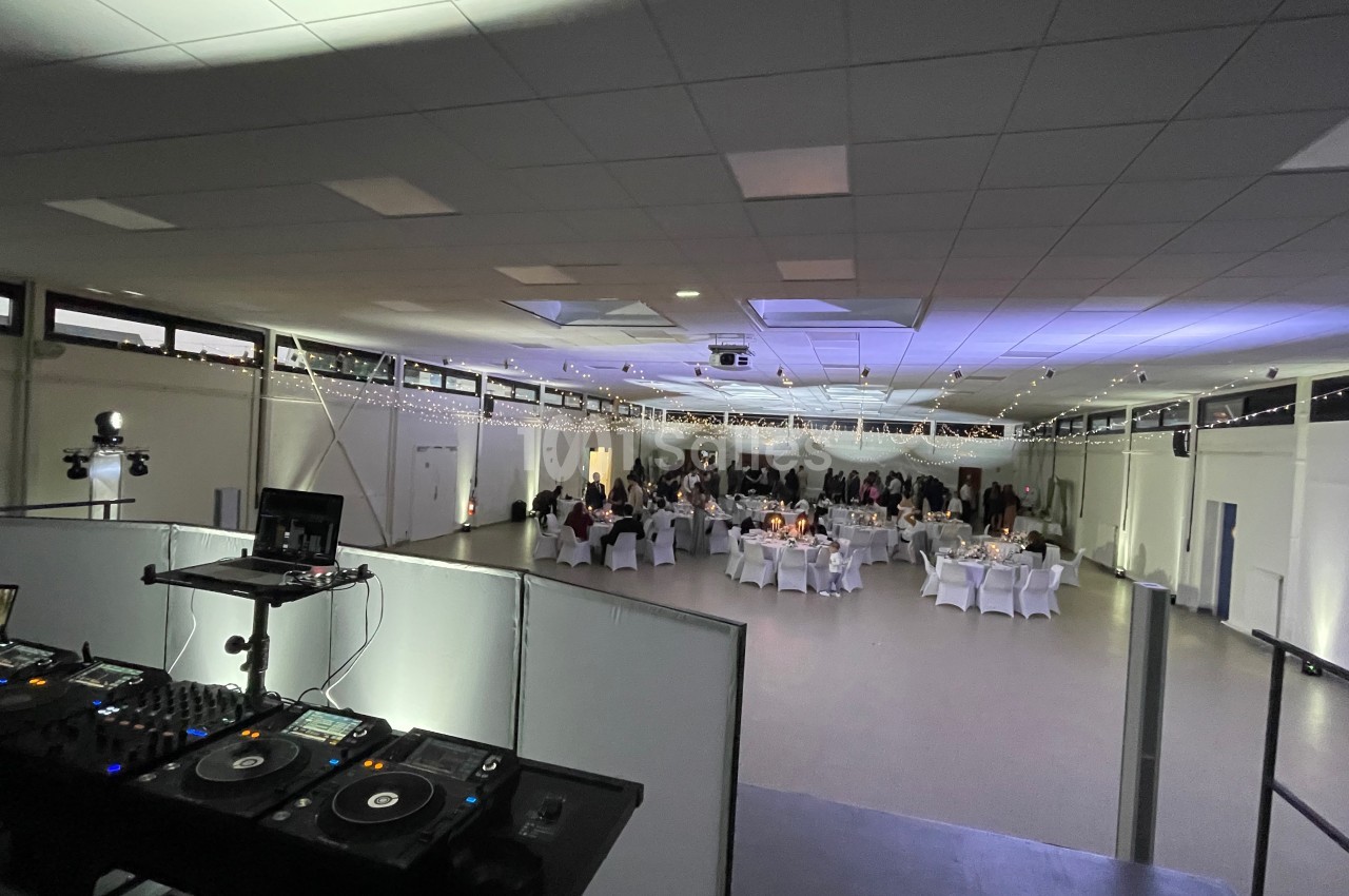 Salle de réception éclairée avec des invités assis à des tables rondes, vue depuis une régie DJ au premier plan.