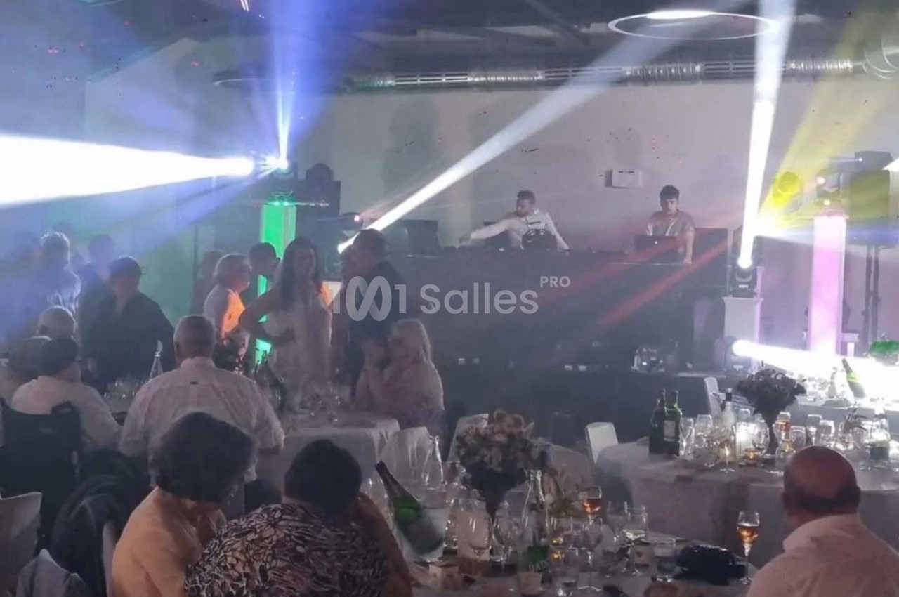 Des convives assis et debout dans une salle de réception éclairée par des jeux de lumière, avec un DJ en arrière-plan.