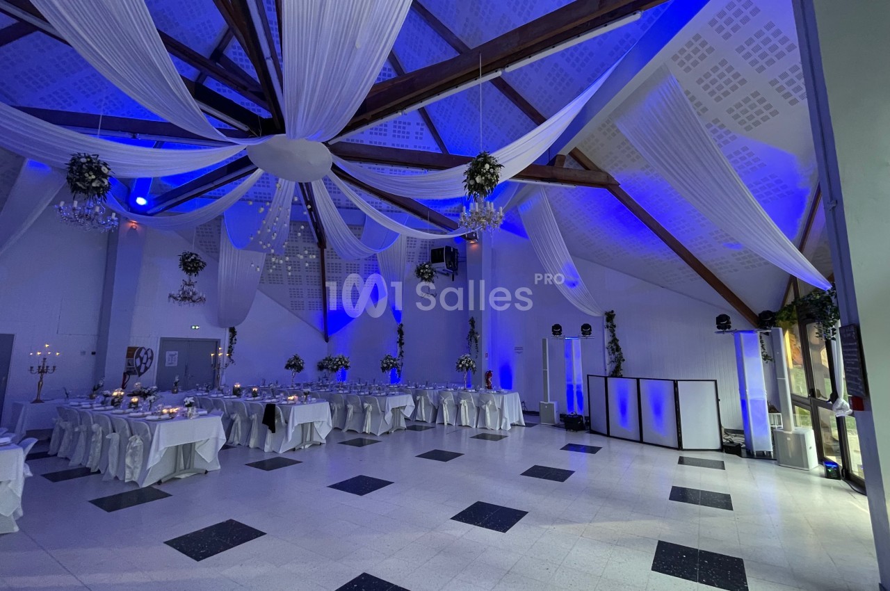 Salle de réception décorée avec des drapés blancs suspendus, éclairage bleu et tables dressées pour un événement.
