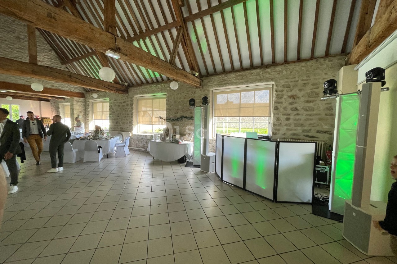 Salle de réception avec poutres apparentes, tables dressées et équipement lumineux installé près des fenêtres.