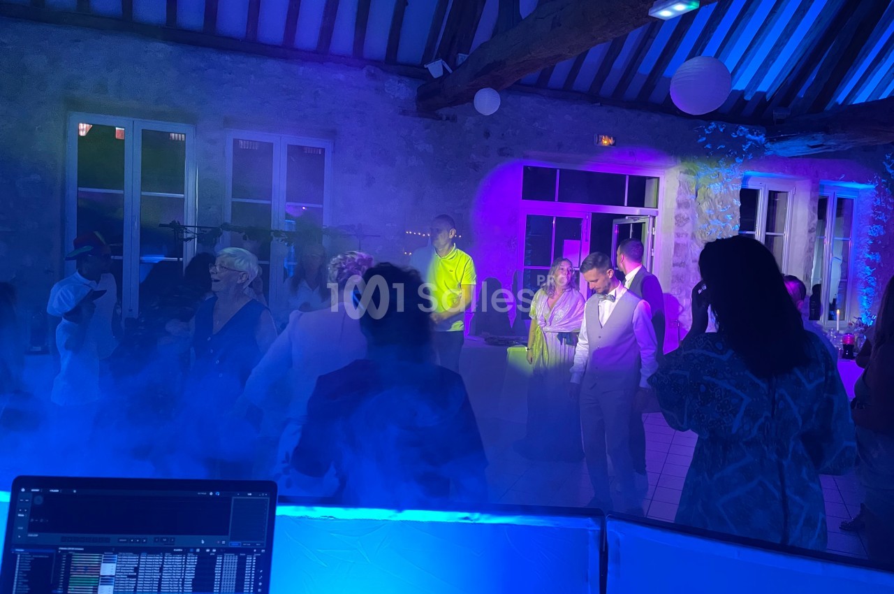 Personnes dansant et discutant dans une salle éclairée par des lumières colorées, avec un DJ en arrière-plan.