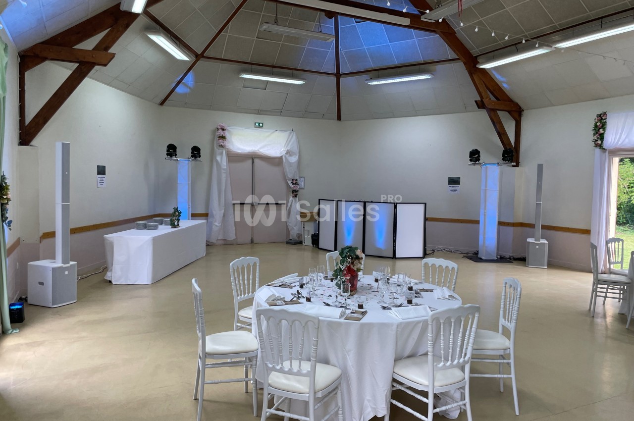 Salle de réception décorée avec des tables dressées, des chaises blanches et un éclairage discret.