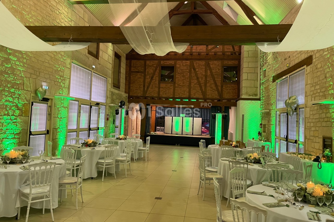 Salle de réception décorée avec des tables rondes dressées, éclairage vert et poutres apparentes en bois.