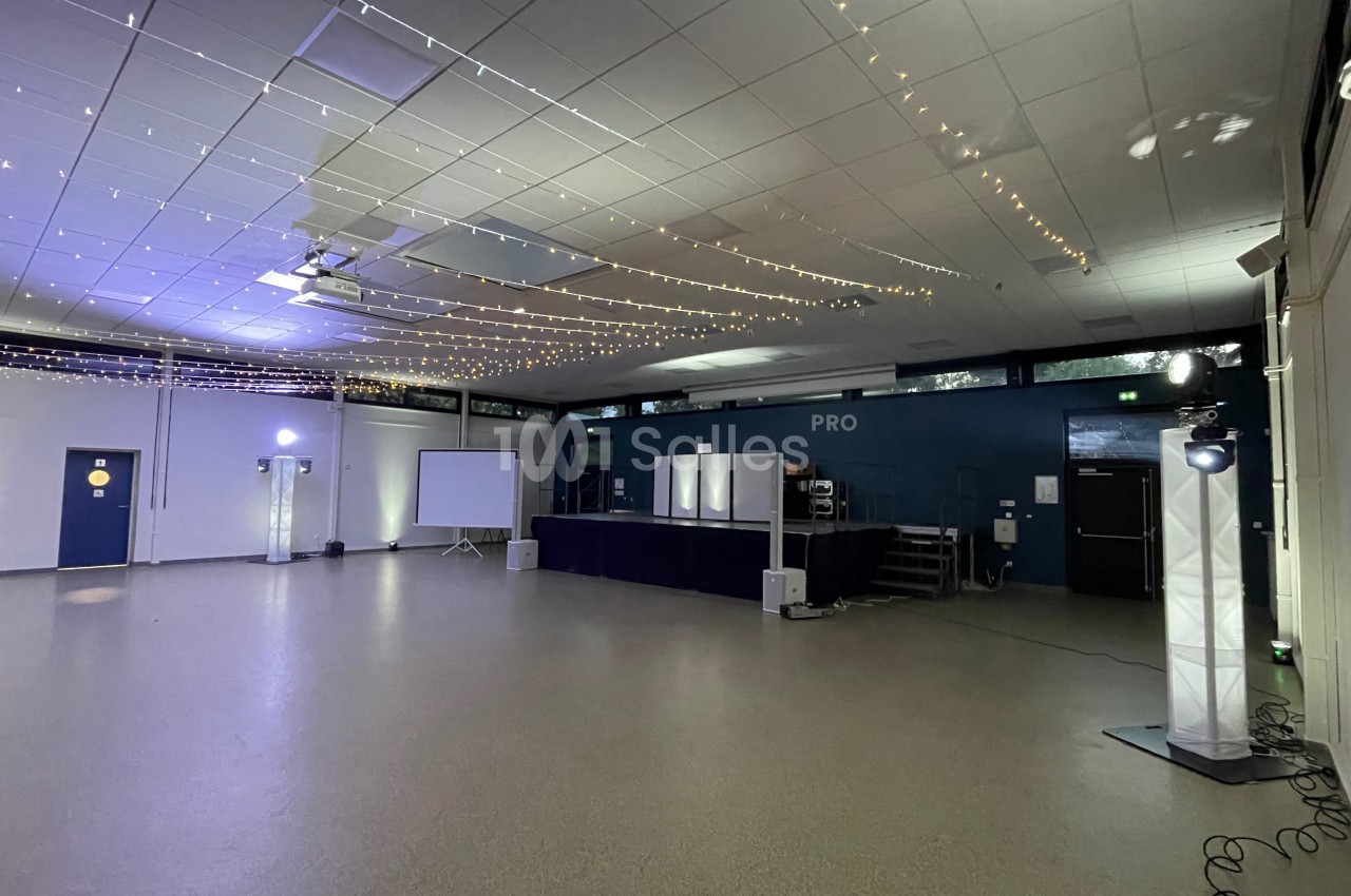 Salle vide avec scène, projecteurs, guirlandes lumineuses au plafond et écran de projection sur le côté.