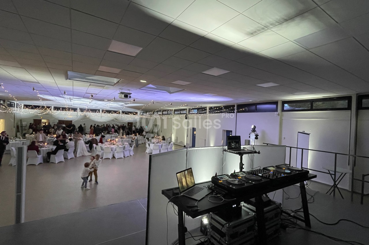 Salle de réception éclairée avec des invités assis à des tables décorées, vue depuis une table de mixage de DJ.
