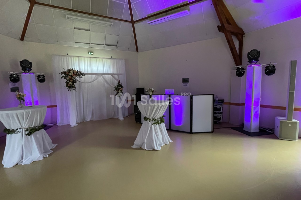 Salle de réception décorée avec des tables hautes drapées, un rideau blanc fleuri et un éclairage violet.