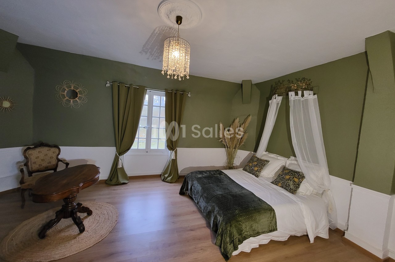 Chambre avec murs verts, lit double à baldaquin, table ronde en bois, fauteuil ancien et fenêtre donnant sur l'extérieur.