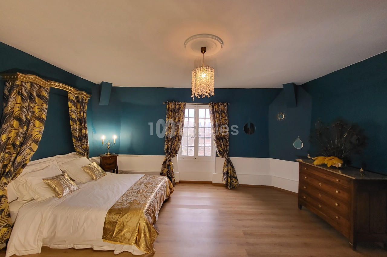 Chambre élégante avec lit à baldaquin, murs bleu profond, parquet clair et lumière naturelle entrant par une fenêtre.