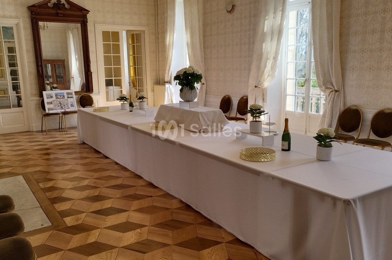 Salle lumineuse avec parquet en bois, tables blanches décorées de plantes et bouteilles, entourées de chaises.