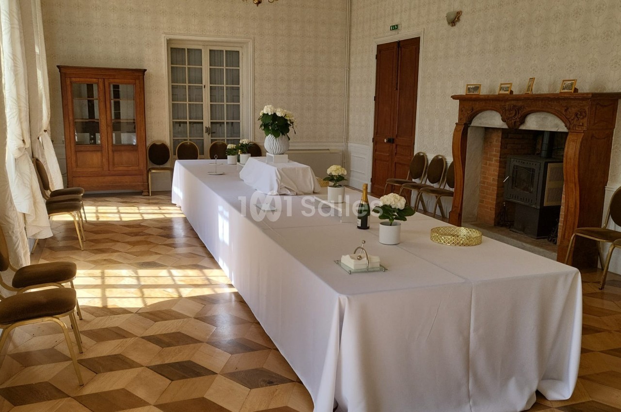 Salle lumineuse avec une grande table rectangulaire recouverte de nappes blanches, décorée de fleurs et entourée de chaises.