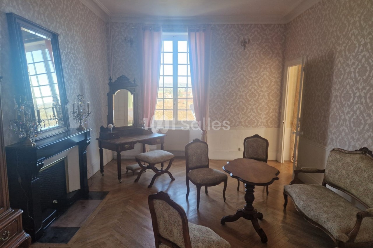 Salon ancien avec parquet en bois, mobilier classique, cheminée, miroir, coiffeuse et grande fenêtre avec rideaux roses.