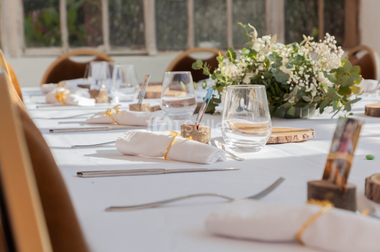 Table dressée avec nappes blanches, verres, couverts, serviettes pliées et décoration florale au centre.