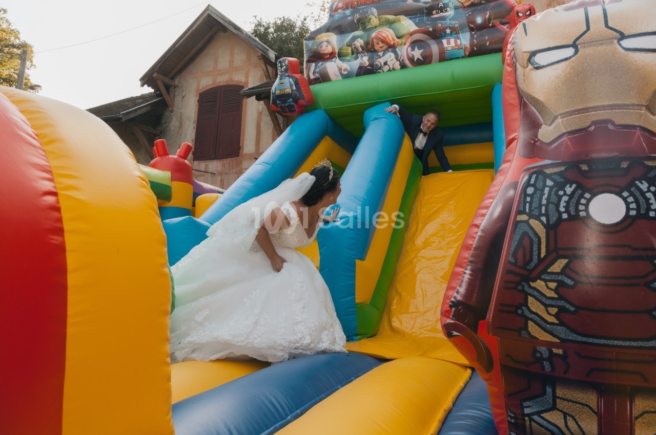Une mariée monte un toboggan gonflable coloré décoré de super-héros, poursuivant un enfant qui glisse.