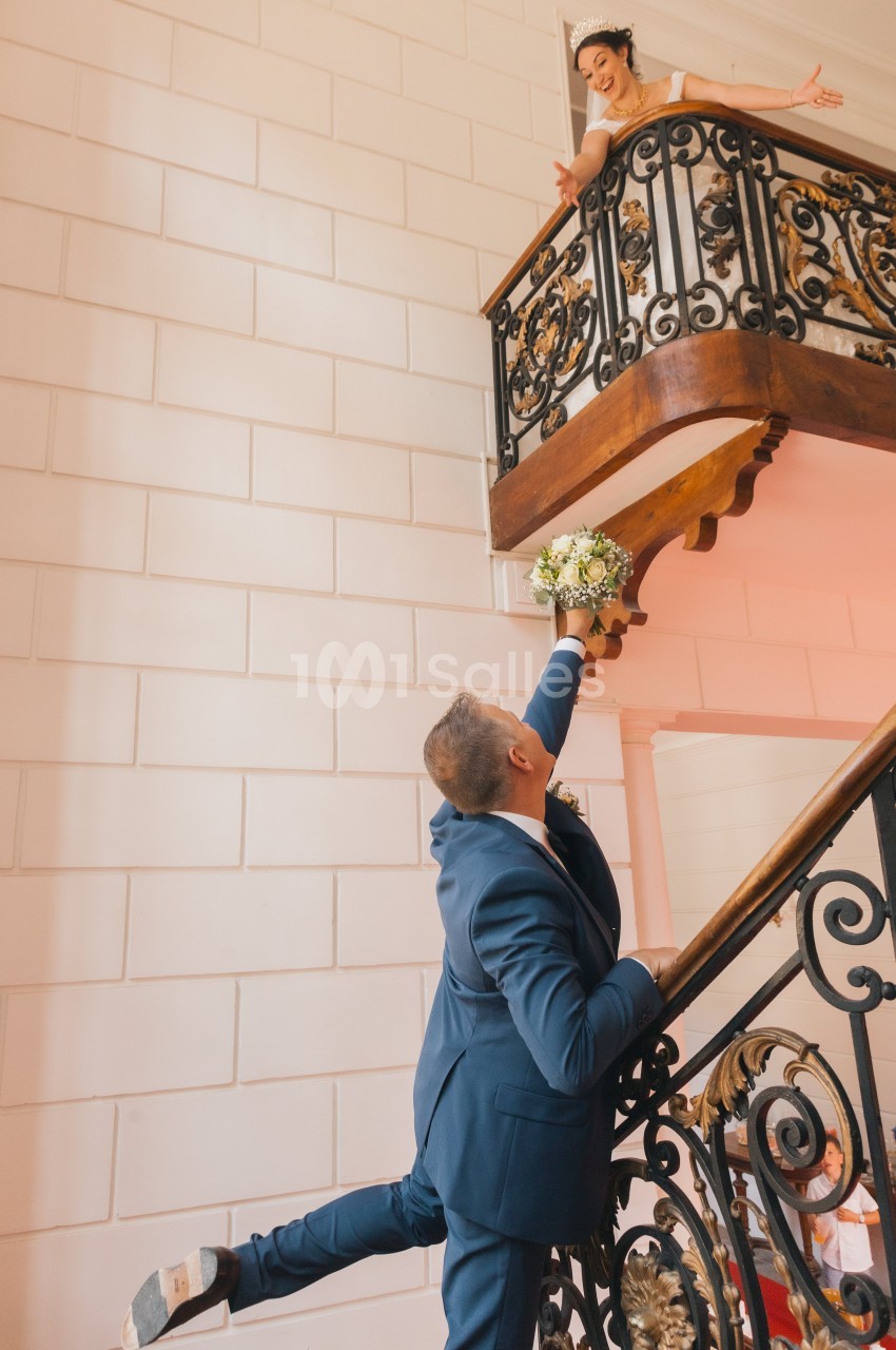 Un homme tend un bouquet de fleurs à une femme souriante sur un balcon, dans un escalier intérieur élégant.
