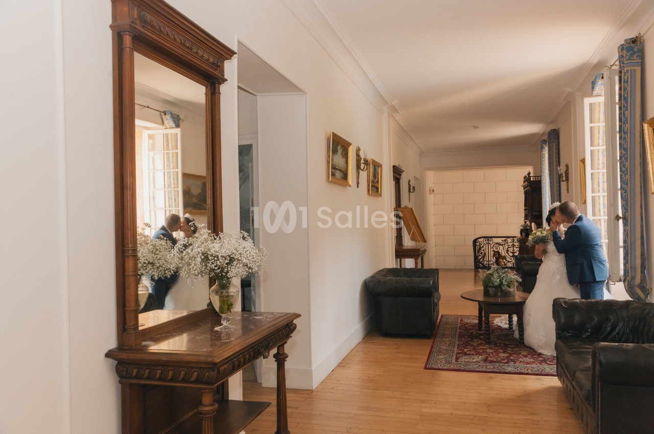 Un couple en tenue de mariage s'enlace dans un salon lumineux avec des meubles anciens et un grand miroir.