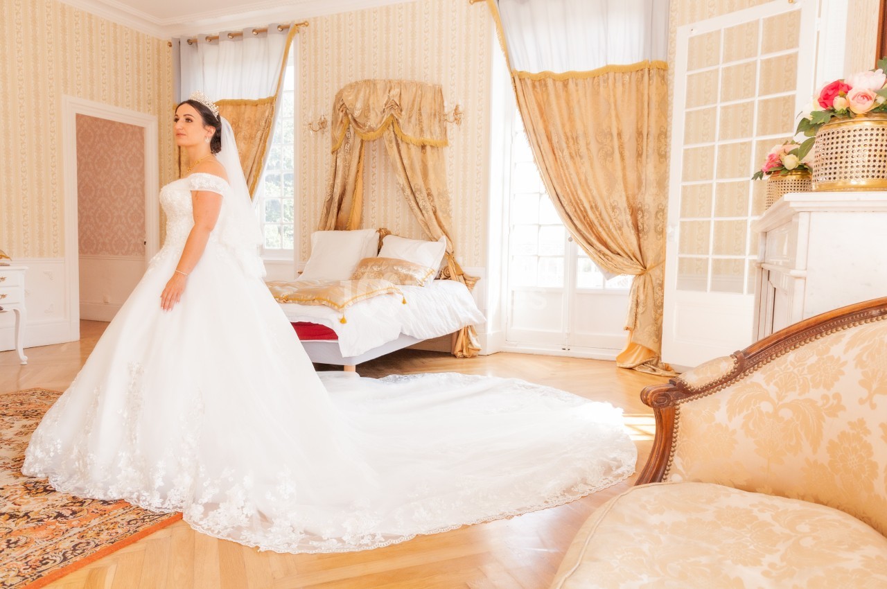 Femme en robe de mariée blanche dans une chambre lumineuse avec lit à baldaquin et rideaux dorés.