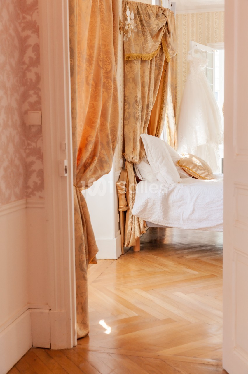 Lit à baldaquin avec rideaux dorés dans une chambre lumineuse, parquet en bois et robe de mariée suspendue en arrière-plan.