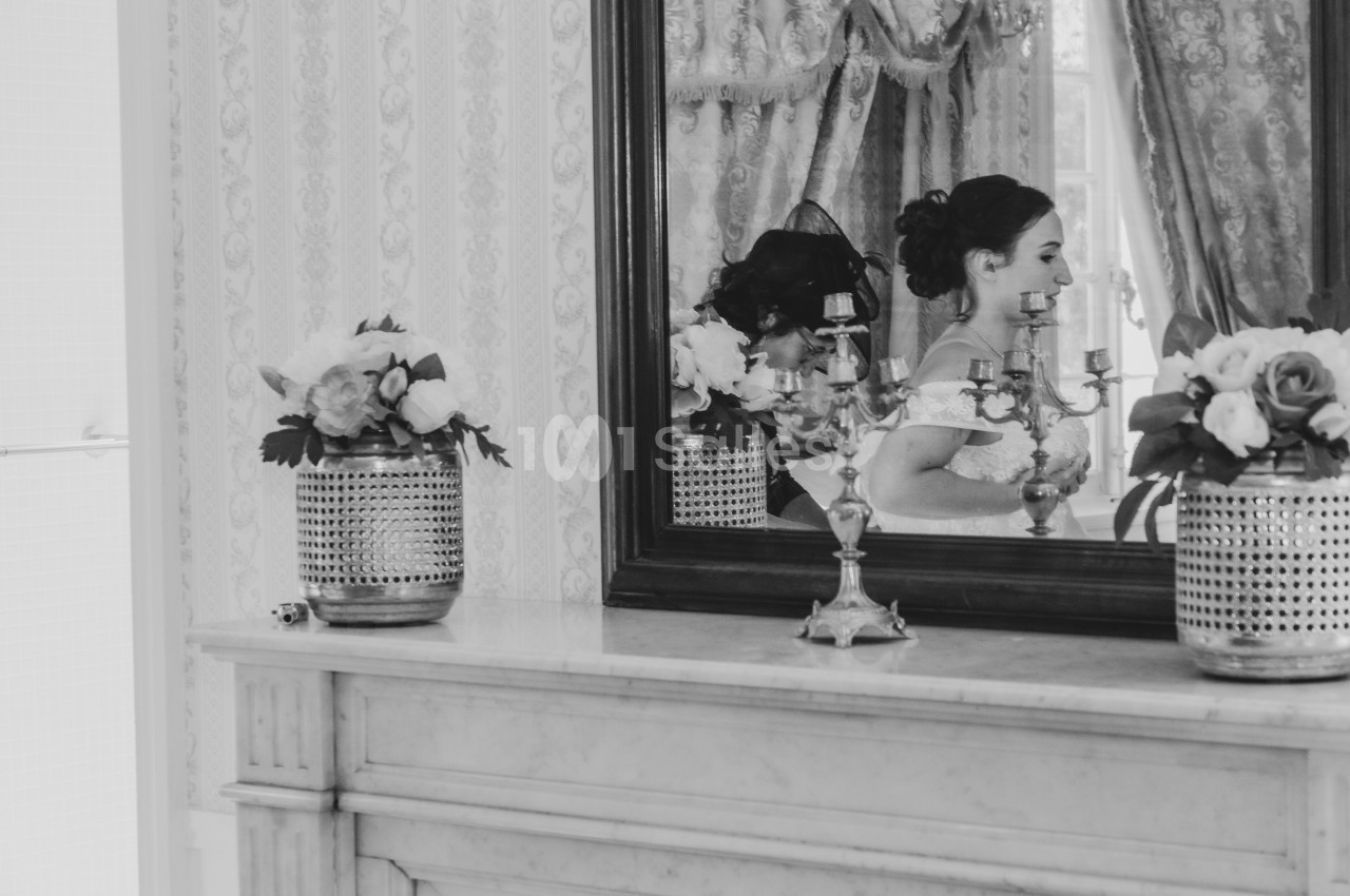 Une femme en robe de mariée se reflète dans un miroir au-dessus d'une cheminée décorée de fleurs et de chandeliers.