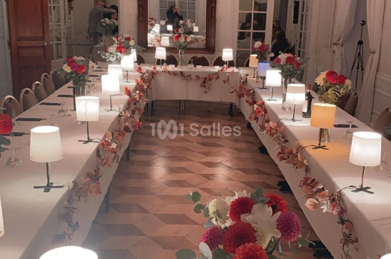 Salle élégante avec tables en U décorées de fleurs rouges et blanches, éclairées par des lampes de table.