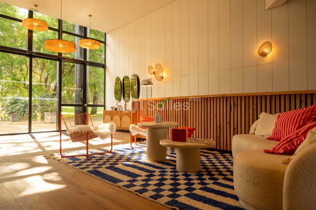 Salon lumineux avec grandes baies vitrées, mobilier moderne, tapis à motifs bleus et blancs, et vue sur un jardin verdoyant.
