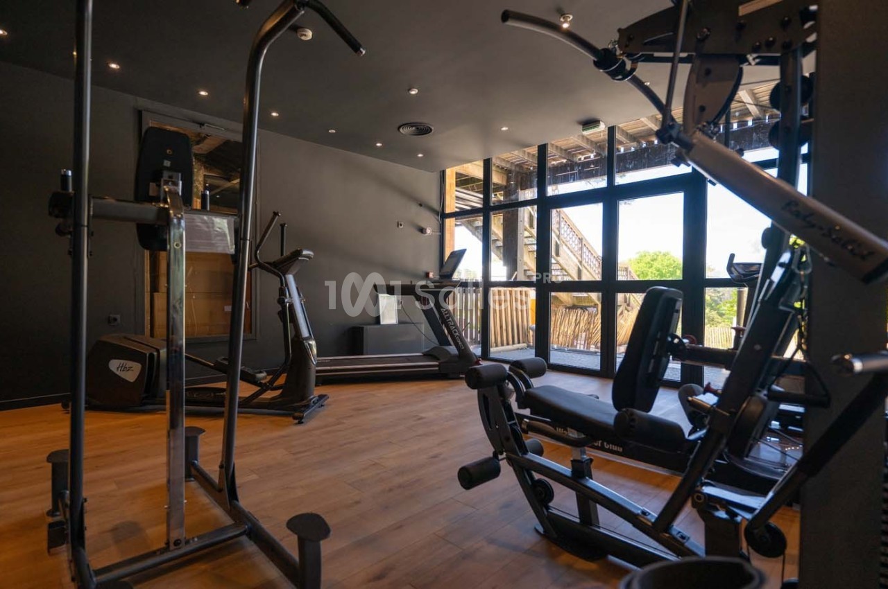 Salle de sport équipée de machines de musculation et de cardio, avec parquet et grandes fenêtres laissant entrer la lumière…