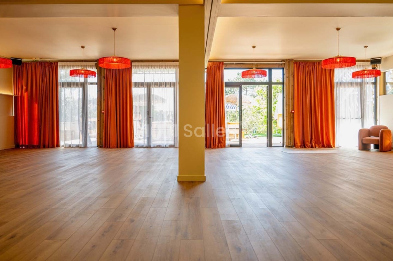 Salle lumineuse avec parquet en bois, grandes baies vitrées, rideaux orange et suspensions rouges.