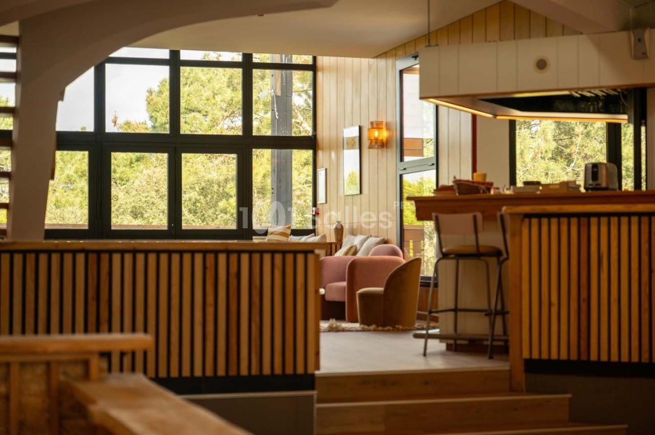 Intérieur lumineux avec grandes fenêtres, mobilier en bois et coin salon cosy avec fauteuils et table basse.